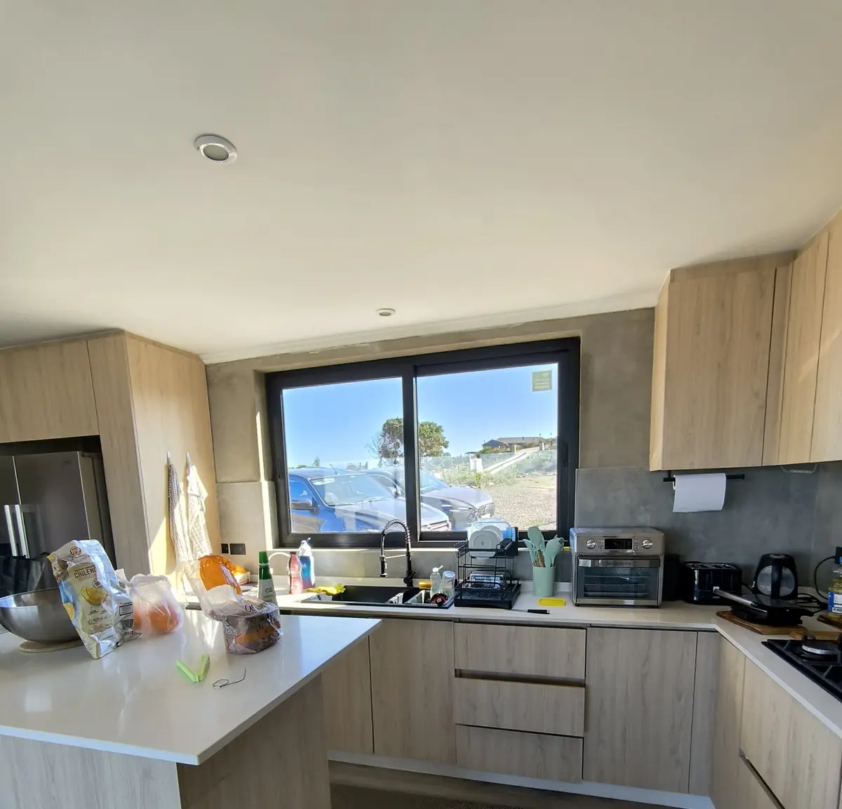 Cocina terminada y en uso: muebles de madera clara, mesón blanco y ventana hacia el exterior