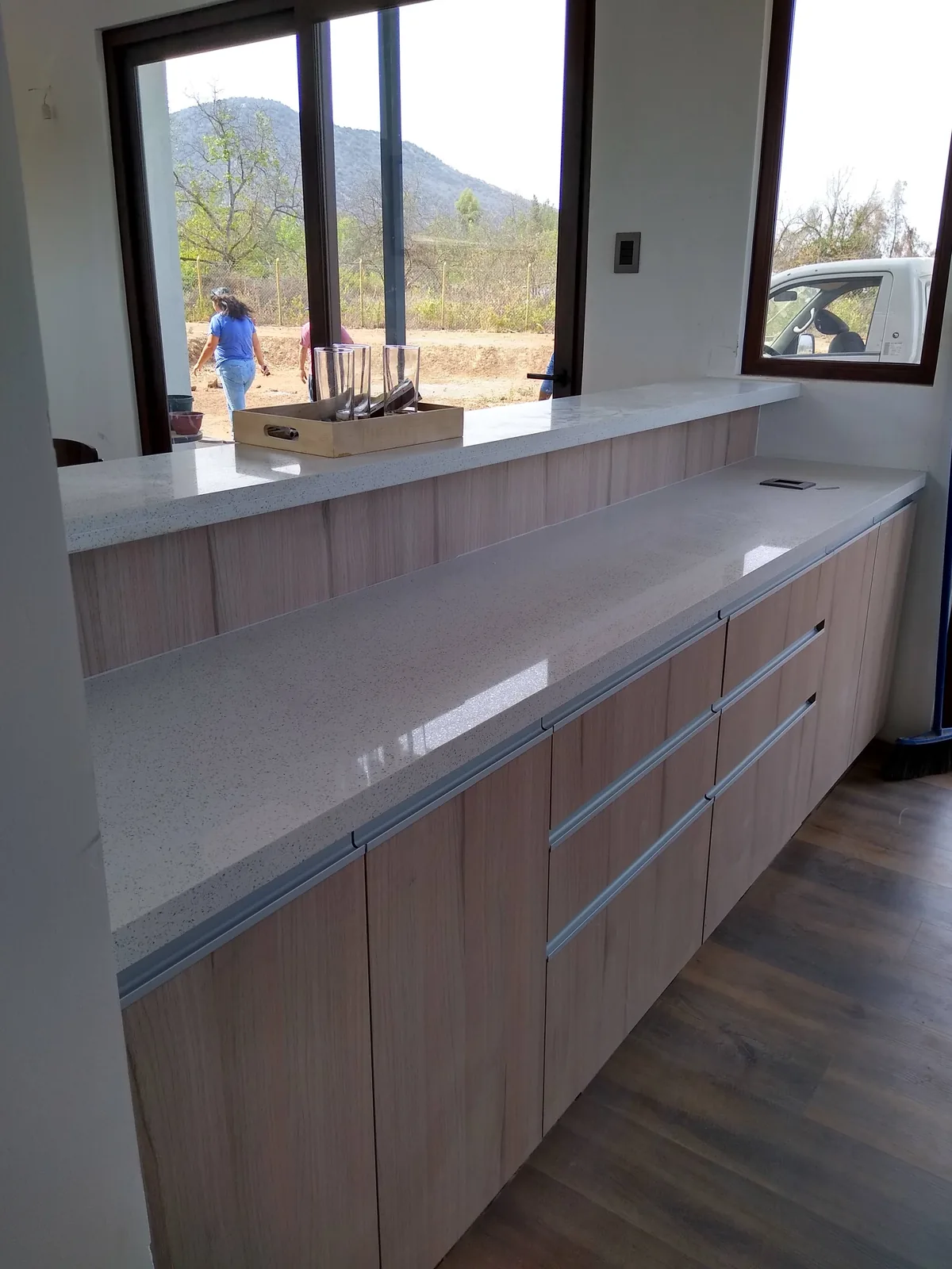Mesón de cocina de cuarzo blanco con muebles de madera clara y ventana con vista al cerro