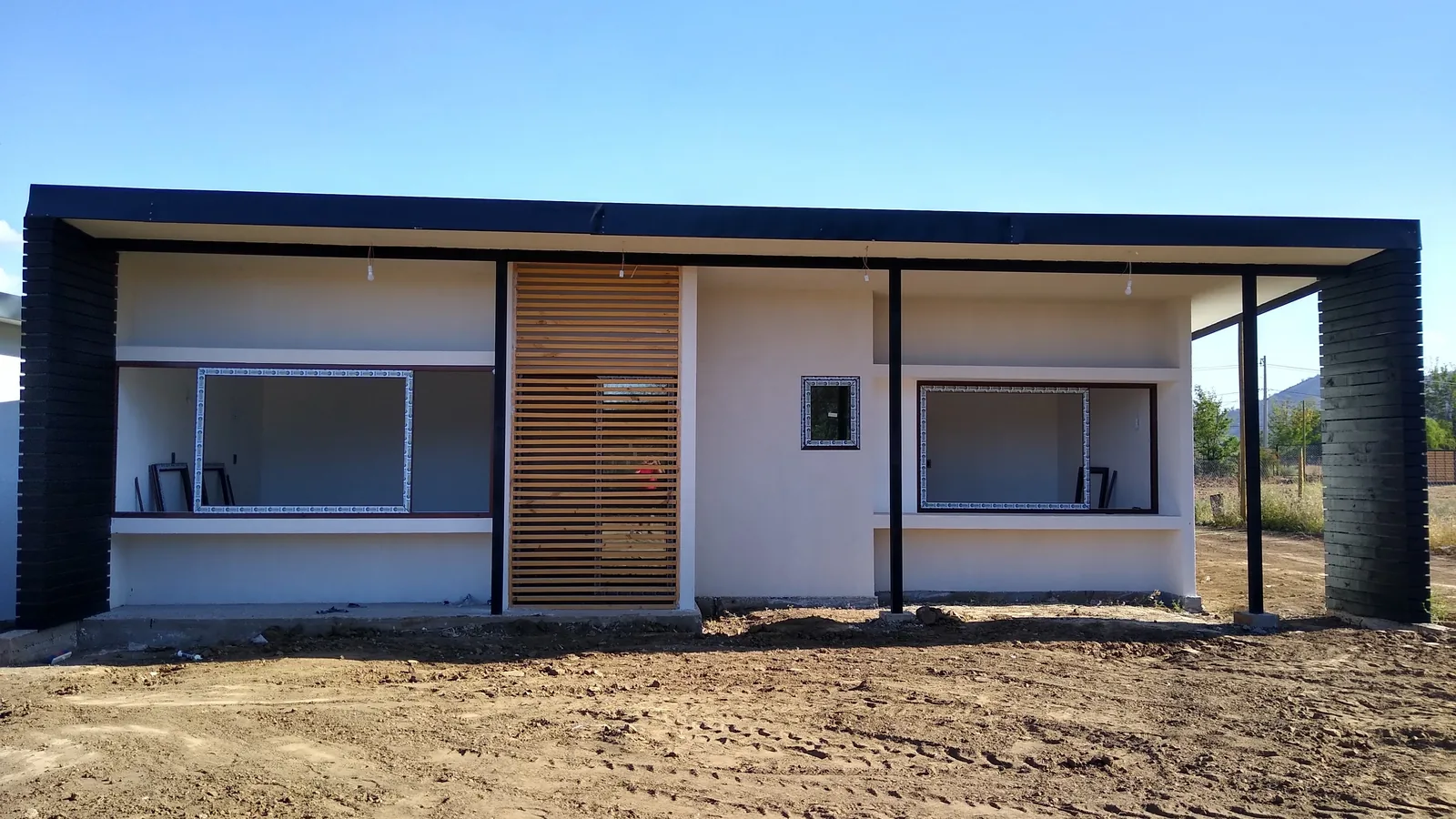 Fachada en terminaciones: alero negro corrido, celosía de madera vertical y ventanales aún protegidos